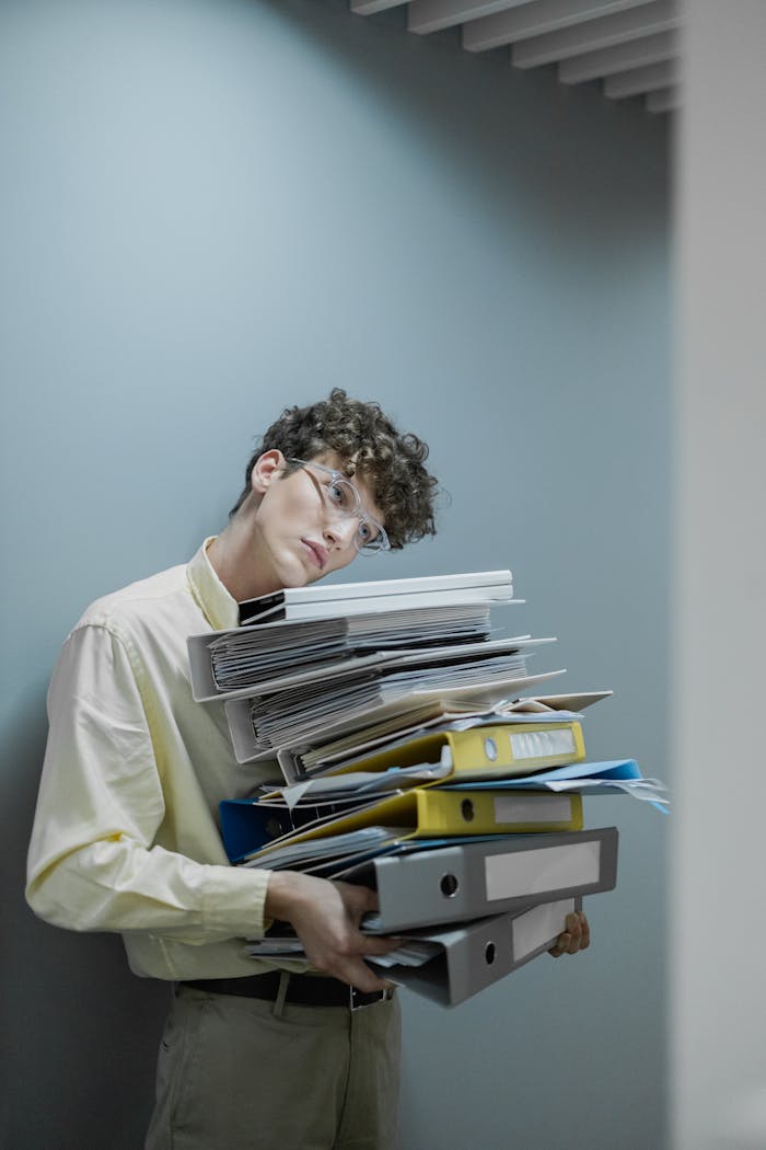 Young man with glasses carrying a stack of files looking tired. Office setting with gray wall.